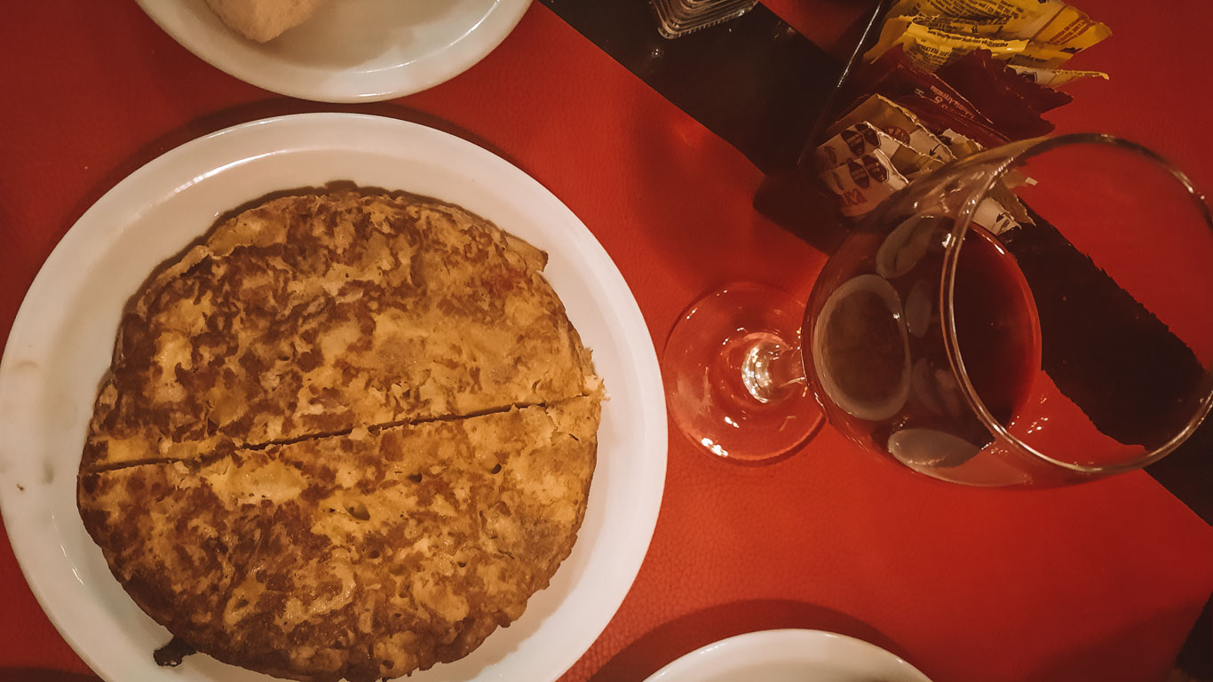 Spanish potato omelette (tortilla de papas) served on a white plate over a red tablecloth, paired with a glass of wine.