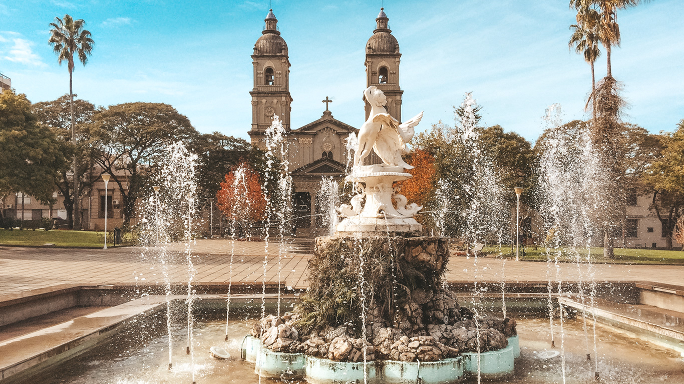 Ornate fountain with angelic statues and water jets in a plaza, framed by palm trees and a twin-towered church in the background under a bright blue sky.