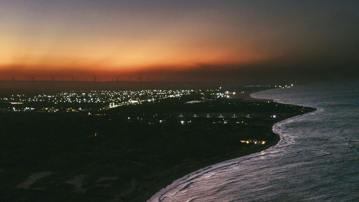 Night view of São Miguel do Gostoso, with city lights twinkling, wind turbines on the horizon, and the coastline glowing as the waves reflect the orange hues of the setting sun.