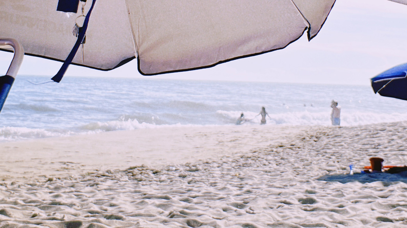 View from under a large beach umbrella showing a sandy shore, children playing in the ocean waves, and sunbathers relaxing by the water.