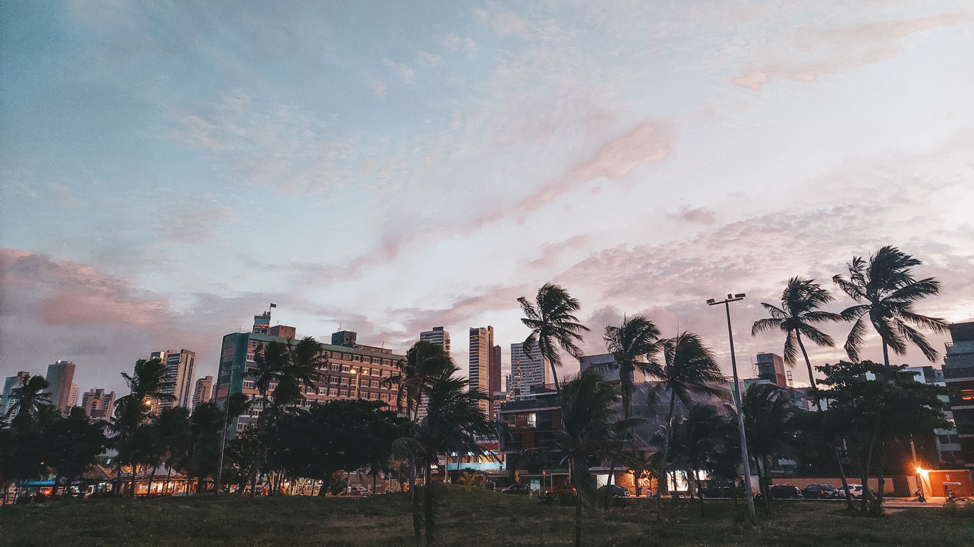 A cityscape of Tambaú at sunset, with tall buildings in the background and palm trees swaying in the wind in the foreground. The sky glows soft shades of pink and blue as dusk falls, making it a great choice for where to stay in João Pessoa.