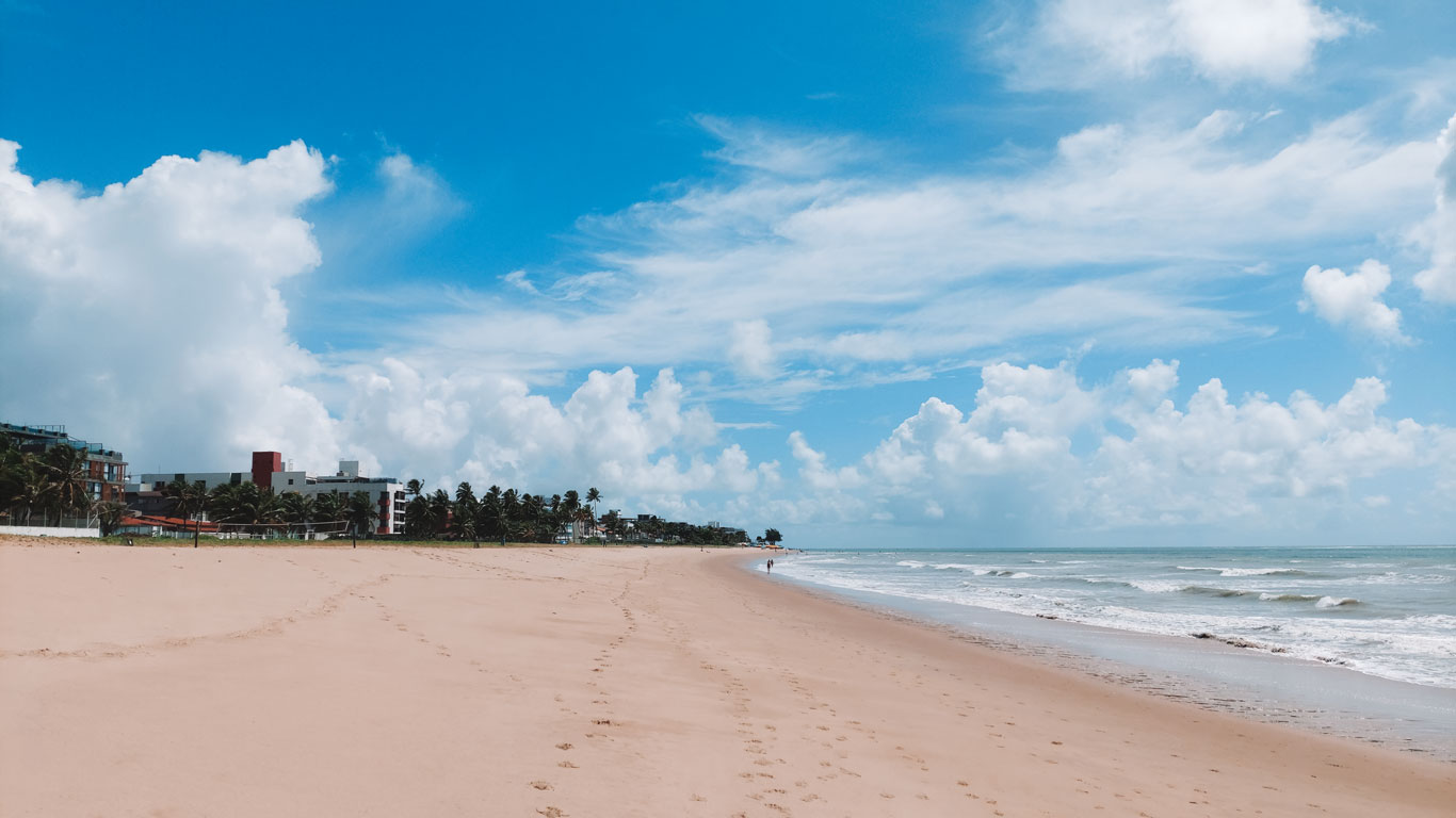 Jardim Oceania Beach, empty of swimmers, with light-colored sand and gentle waves rolling under a blue sky filled with billowy clouds. Low-rise buildings and palm trees line the left side, in João Pessoa.