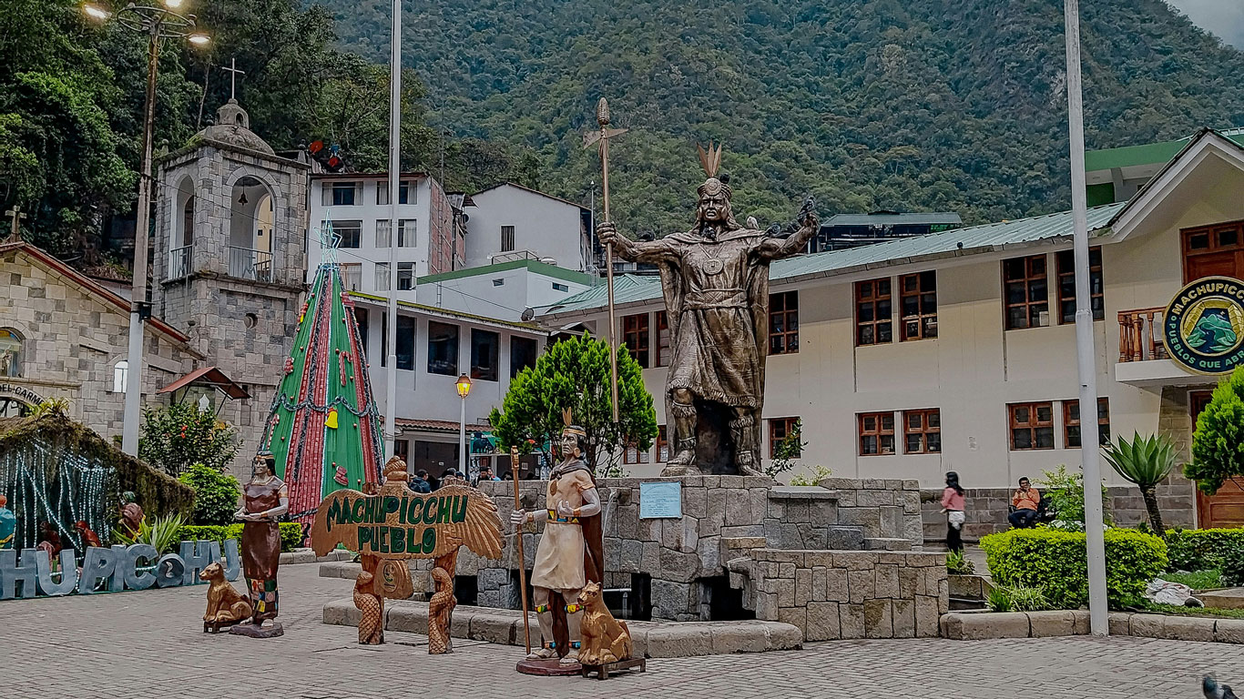 Central plaza in Aguas Calientes, also known as Machu Picchu Pueblo, featuring a large Inca statue, festive decorations, and nearby buildings.