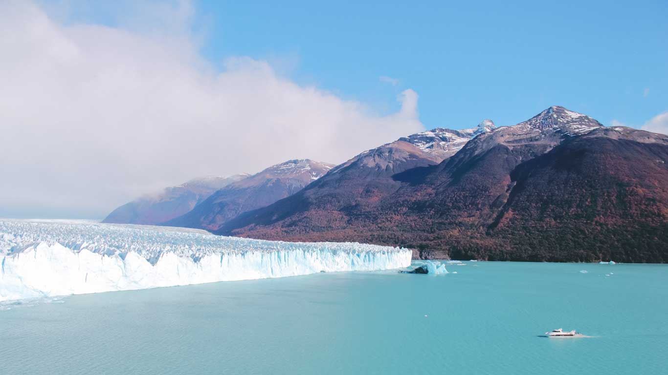 A panoramic view of the Perito Moreno Glacier in Argentina, where bright turquoise water meets a massive wall of blue ice with rugged, snow-capped mountains in the background. A small boat is visible near the glacier, highlighting its immense scale. Perito Moreno Glacier day trip – boat tour in El Calafate.