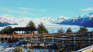 A scenic overlook with wooden walkways and a covered shelter facing the vast Perito Moreno Glacier and the surrounding Andes mountains. Snow-dusted ground and green pine trees frame the viewpoint.