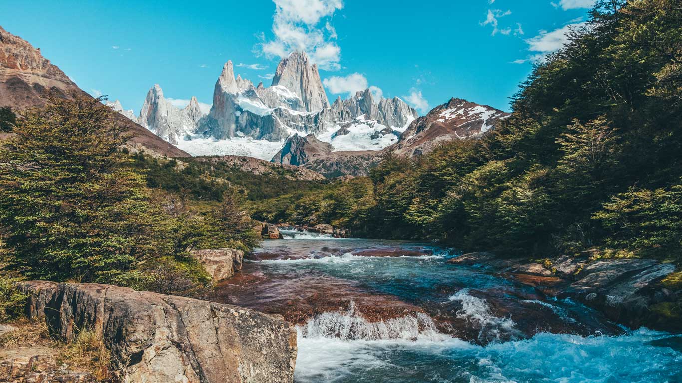 A river flows through a lush green valley toward the snow-covered peaks of Mount Fitz Roy under a bright blue sky. This iconic Patagonia hike is a top choice on trekking tours in El Calafate.