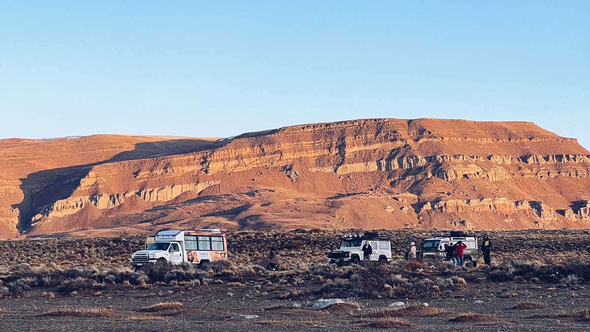 Off-road tour vehicles parked in the Patagonian steppe near layered red rock formations during golden hour. Adventure tours in El Calafate often include 4x4 excursions through this rugged landscape.