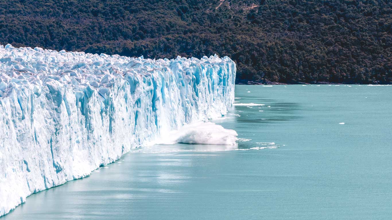 A close-up shot of the towering icy wall of the Perito Moreno Glacier as a chunk of ice dramatically breaks off and splashes into the blue water below, illustrating the process of glacier calving.